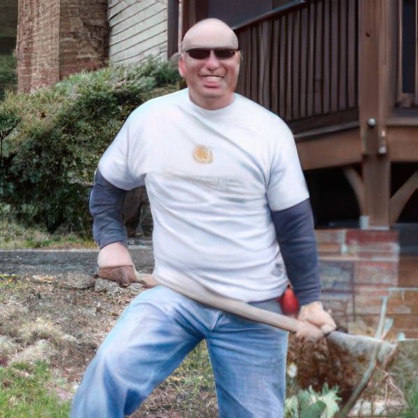 Bald man smiles, holds hoe. Wears white t-shirt, blue jeans, and sunglasses. Working outside in front of a building.