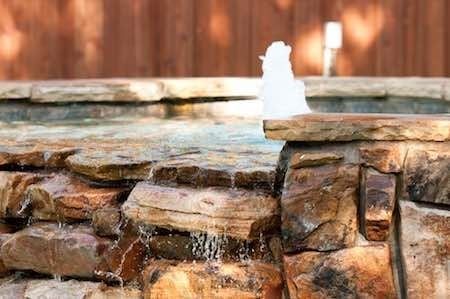 Stone-lined water fountain with water flowing over layered rocks, set against a wooden fence.