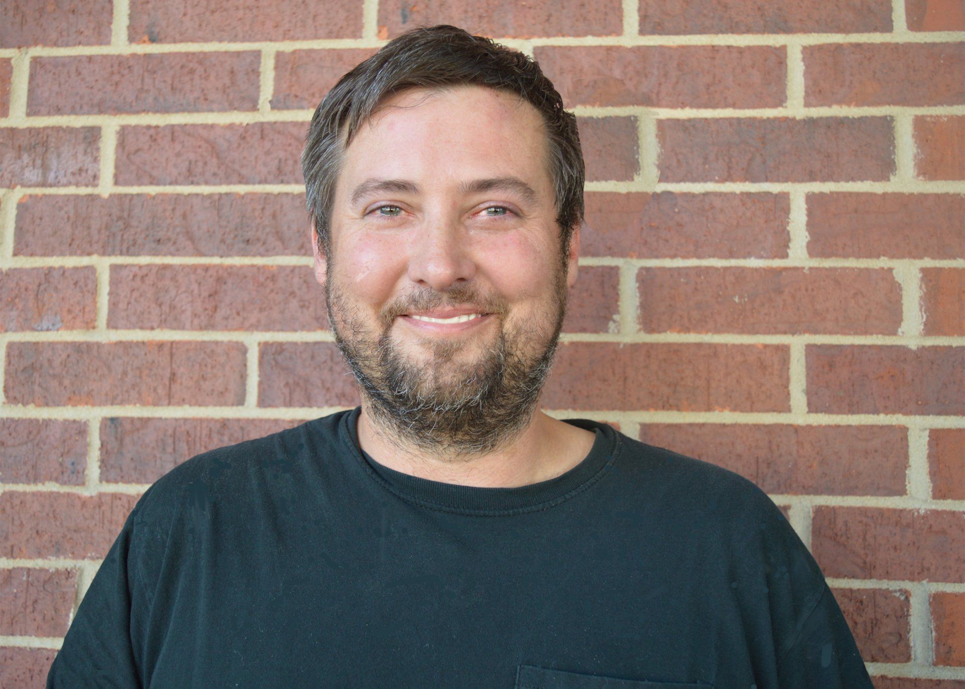 Man with beard smiling in front of a brick wall, wearing a black shirt.