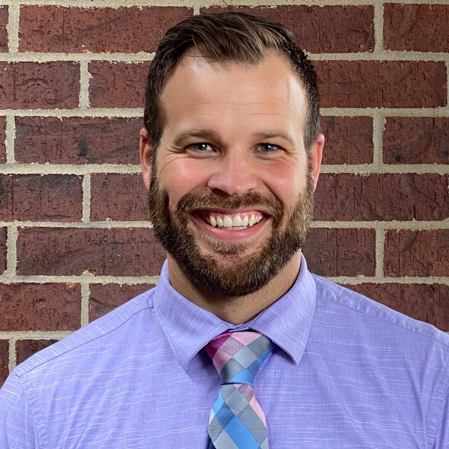Man with a beard smiling in front of a brick wall, wearing a light purple shirt and patterned tie.