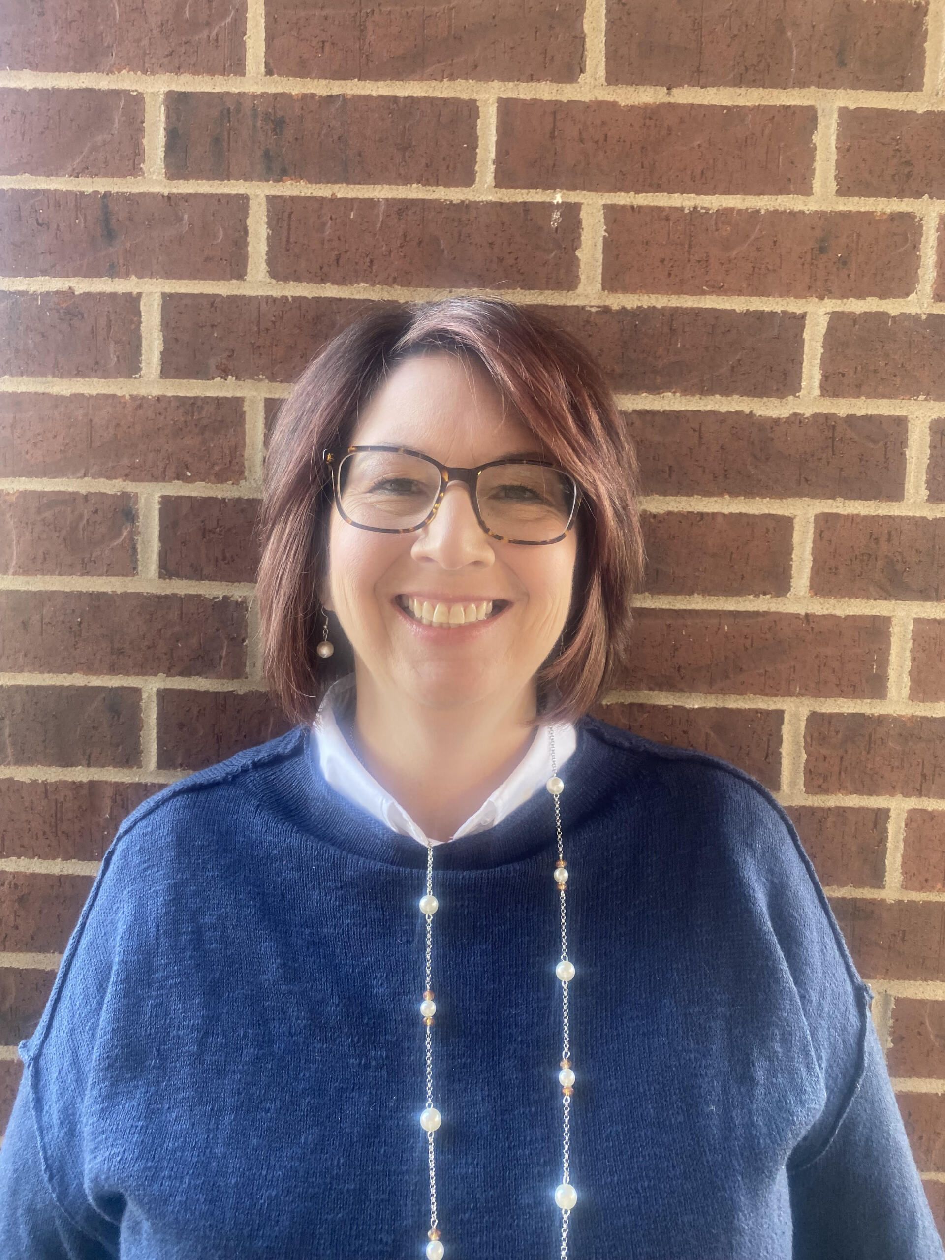 Woman with glasses smiles in front of a brick wall, wearing a blue sweater and pearl necklace.
