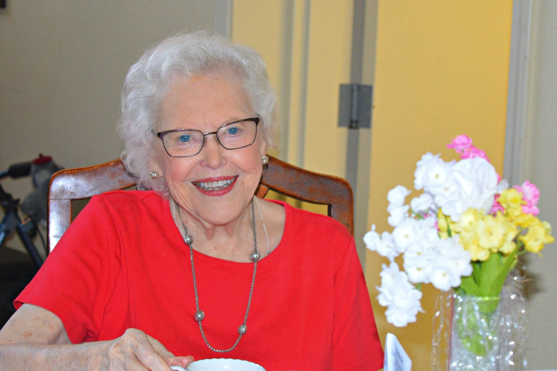 Smiling woman with glasses in red shirt, sitting at a table near a vase of flowers.