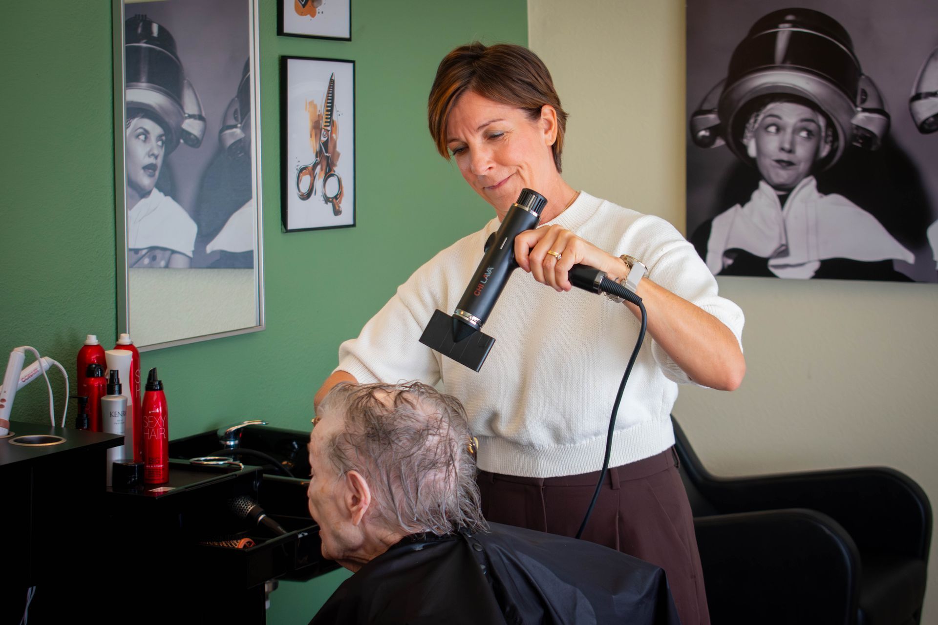 Hairdresser blow-drying a client's wet hair in a salon. Black and white photos of people with hair dryers on the wall.