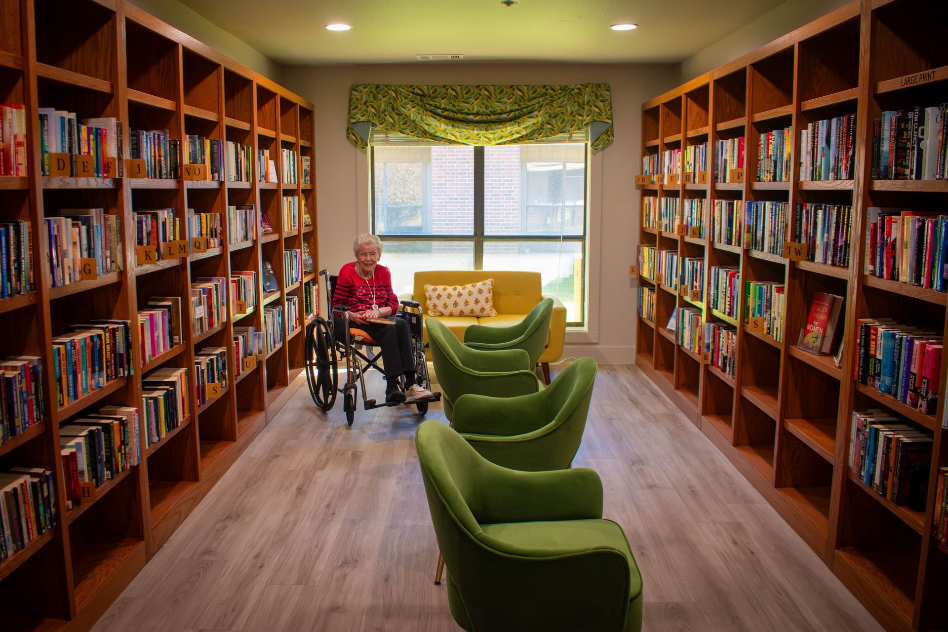 Elderly person in a wheelchair in a library surrounded by bookshelves, sitting near a window and green chairs.