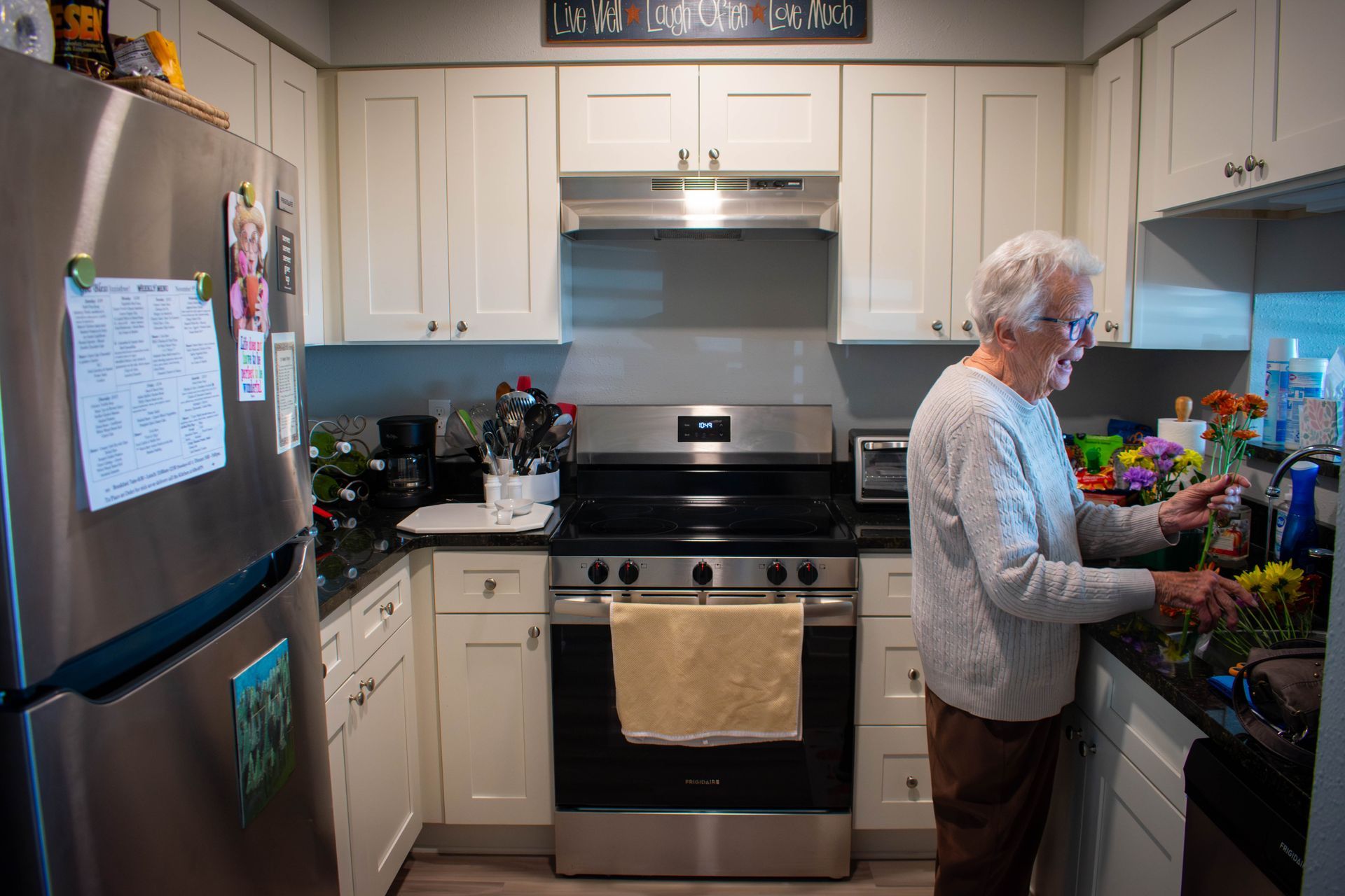 Woman in a kitchen, arranging flowers on a countertop next to a stove. White cabinets, stainless steel appliances.