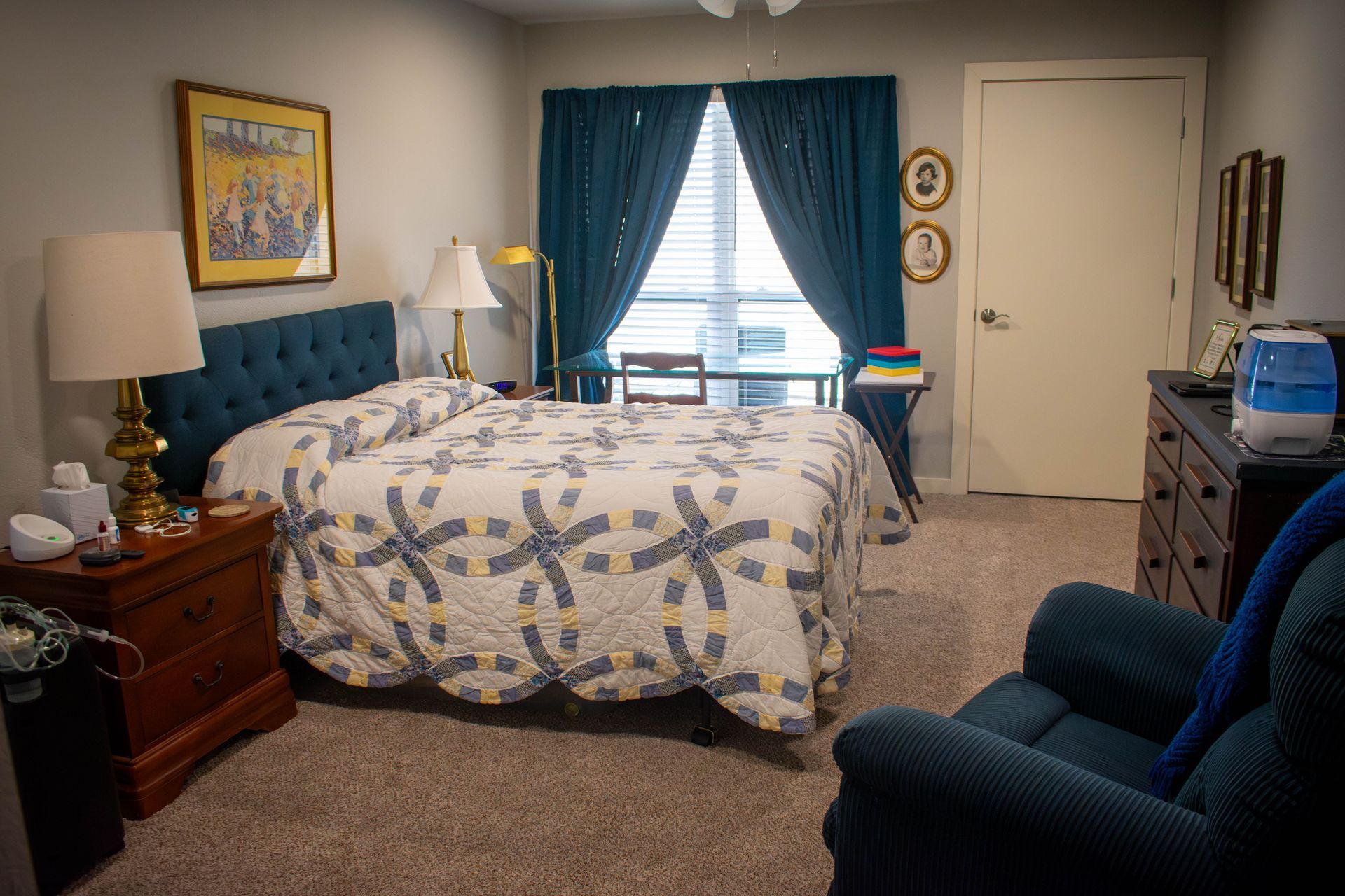 Bedroom with bed, blue quilt, armchair, desk, and teal curtains.