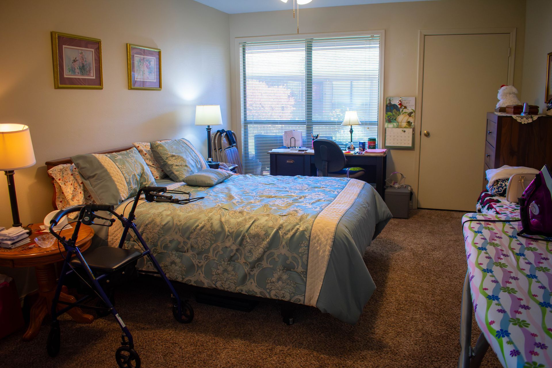 Bedroom with bed, desk, walker, lamp, window with blinds.