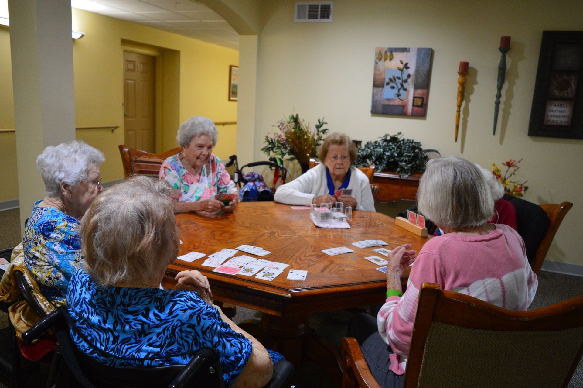 Elderly women playing cards around a wooden table in a well-lit room.