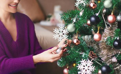 Person in a purple sweater decorating a Christmas tree with white snowflake ornaments and pink baubles