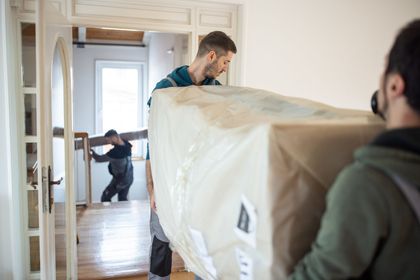Two movers carrying a large wrapped sofa through a narrow hallway while a third person helps in the background