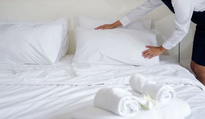 Person making a bed with white linens and rolled towels in a bright room