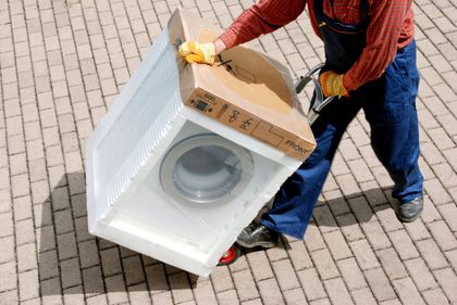 Person carrying a large boxed appliance on a wheeled dolly over a brick-paved surface