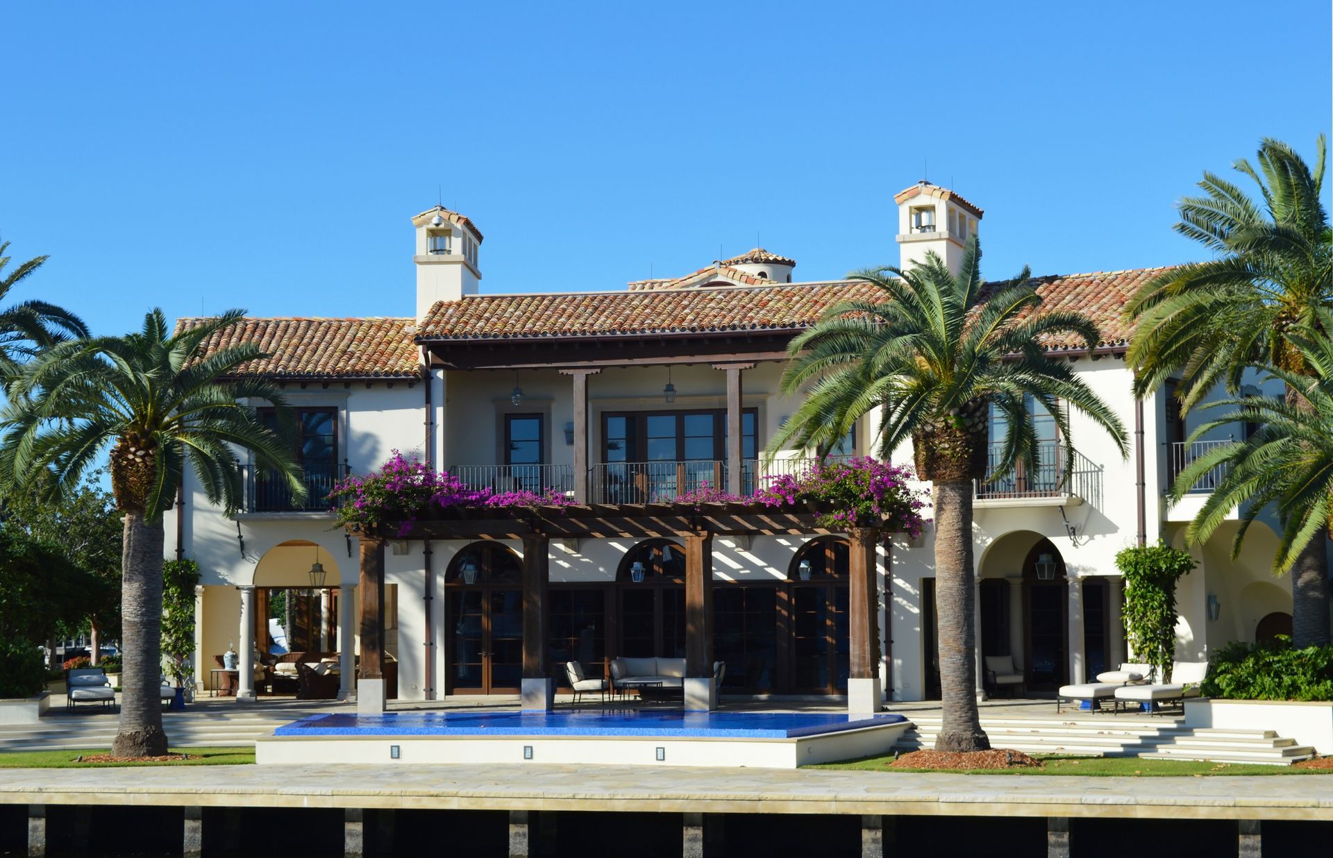 Large Mediterranean-style villa with red-tiled roof, palm trees, and a swimming pool under a clear blue sky