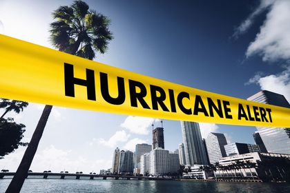 Yellow “HURRICANE ALERT” banner over a coastal city skyline and palm tree under a stormy sky