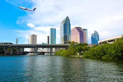 City skyline with a bridge over water and an airplane flying overhead under a blue sky
