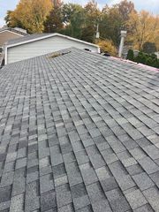 Gray asphalt shingles on a sloped residential roof with a white garage and autumn trees in the background.