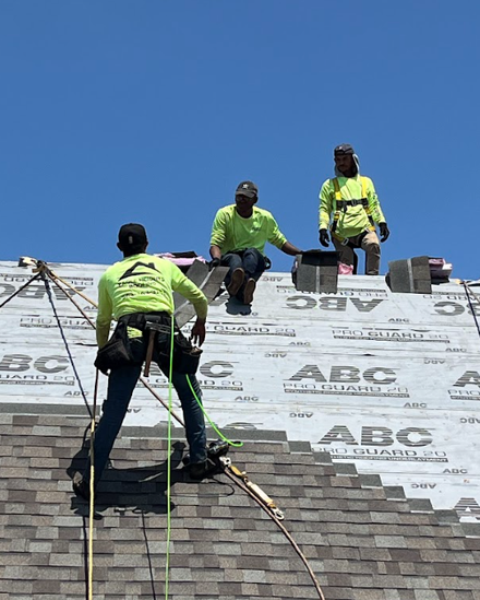 Three workers in neon green safety shirts install shingles on a roof under a clear blue sky.