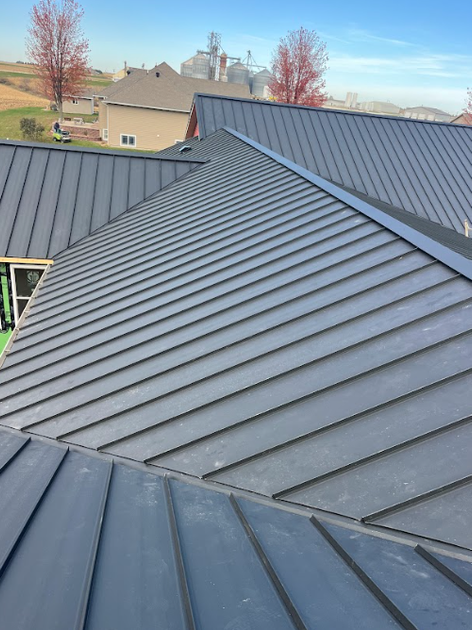 An elevated view of a gray metal roof with standing seams, featuring multiple pitches and a house in the background.