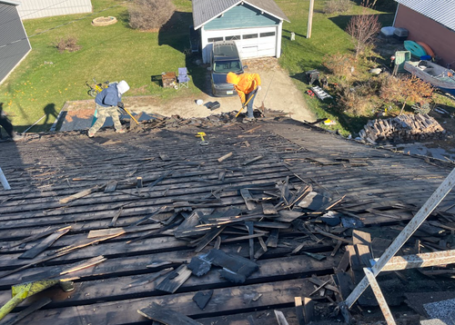 Two people removing old shingles from a house roof, exposing the wood slats underneath in a sunny outdoor setting.