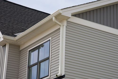 Gray vinyl siding and a dark shingled roof on the corner of a house with white trim, gutters, and a downspout.