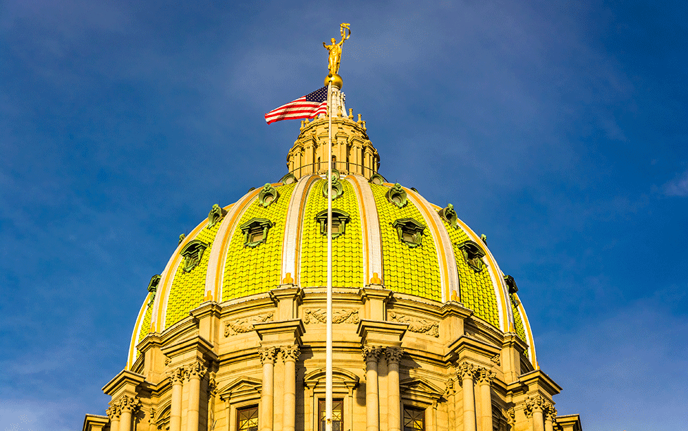 The green, domed roof of the Pennsylvania State Capitol building with an American flag flying against a blue sky.