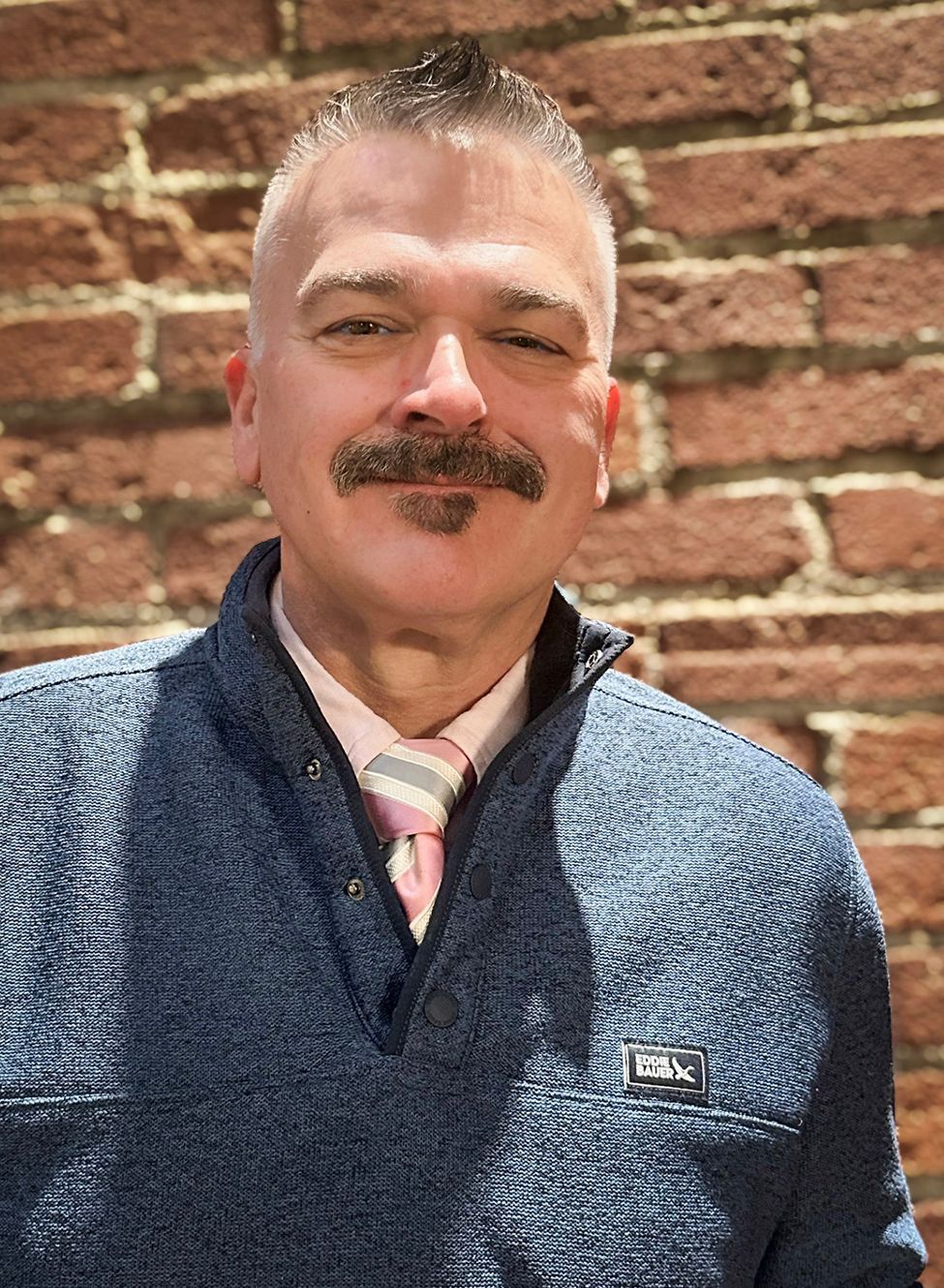 A person with a mustache and goatee wears a blue textured quarter-zip sweater and a striped tie, posing before a brick wall.