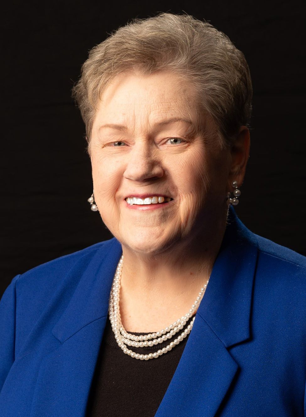 A woman with short hair smiling, wearing a blue blazer and a three-strand pearl necklace against a black background.