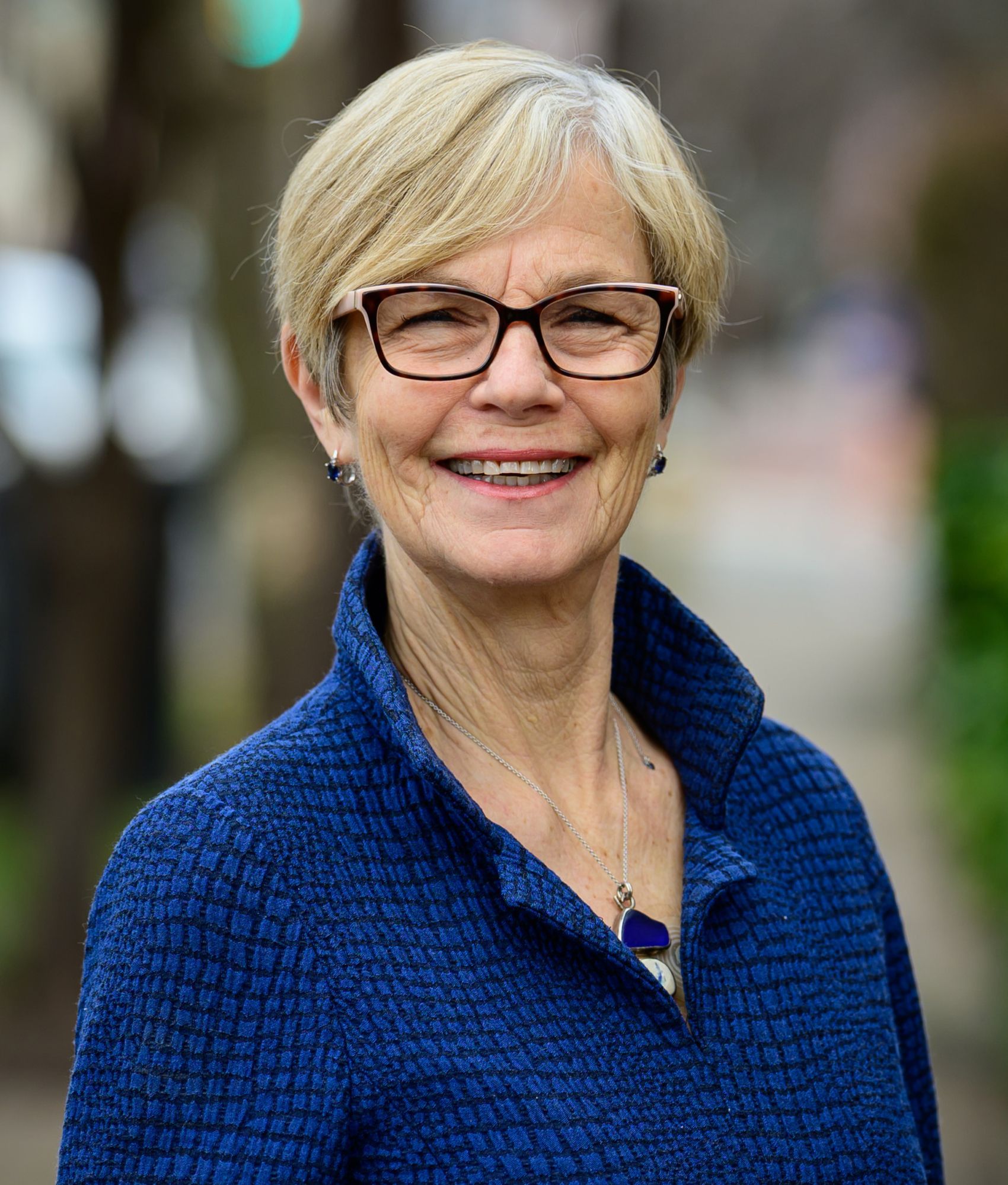Keynote Presenter Name Smiling person in glasses and a blue sweater outdoors with a blurred background