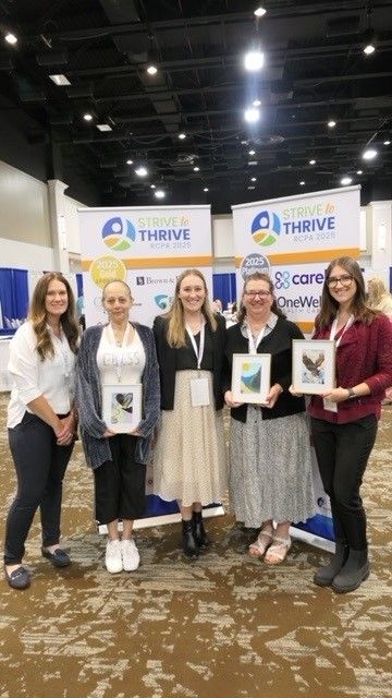 Five people pose in a convention hall in front of banners, holding awards and certificates.