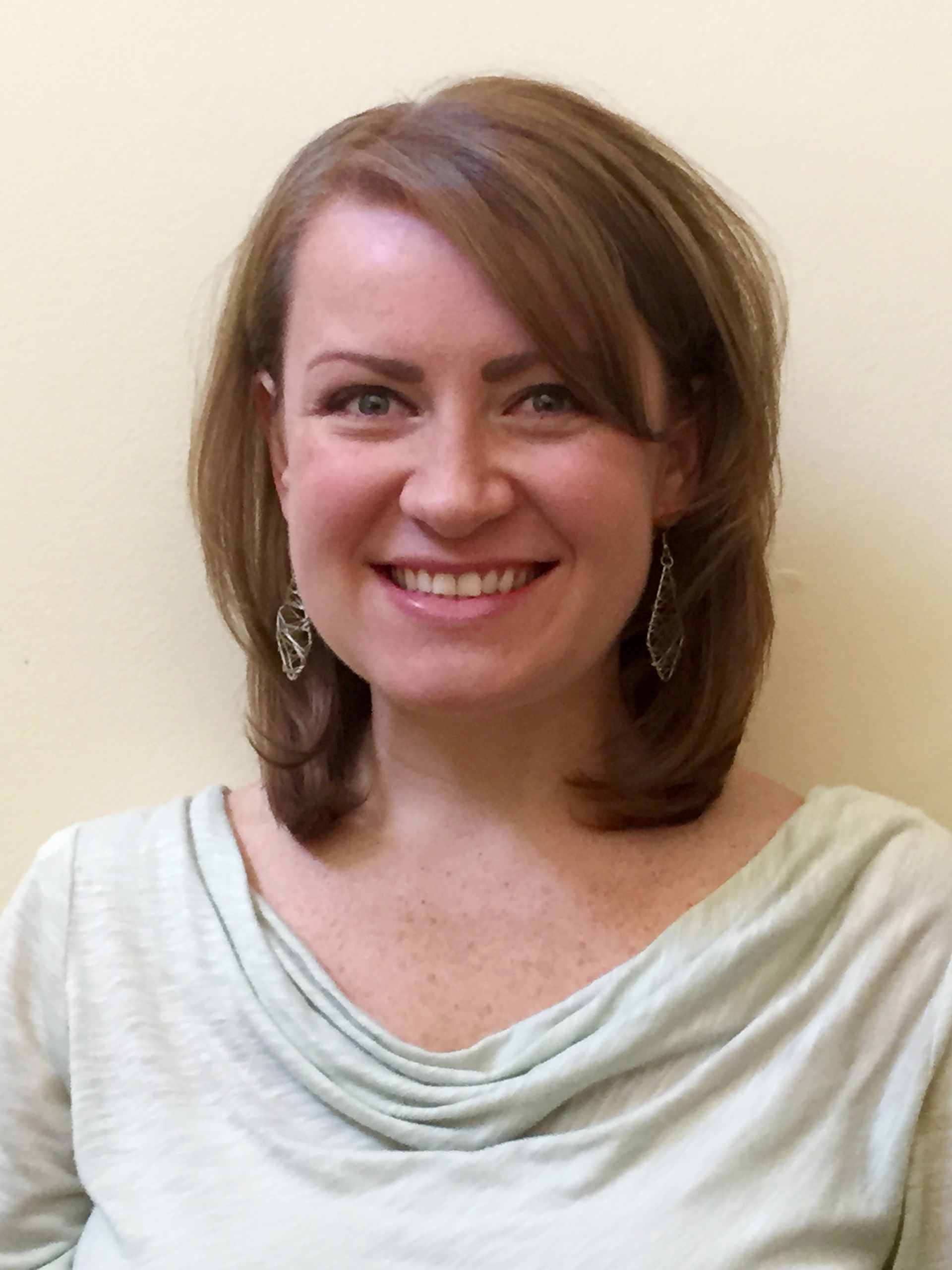 A person with shoulder-length brown hair and dangling earrings smiles warmly against a plain, light background.