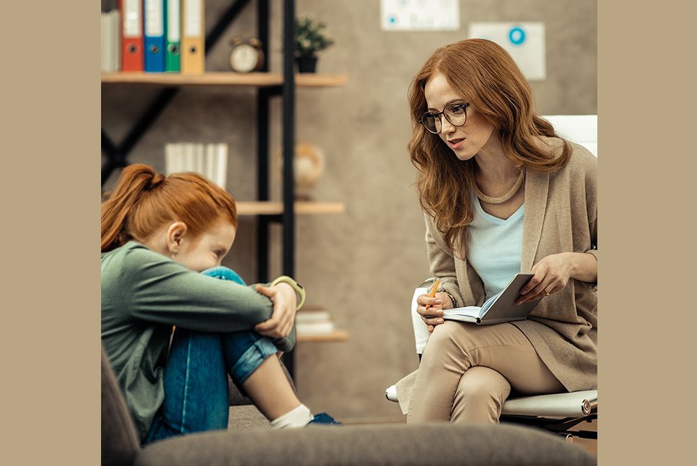 Two women talking in a cozy room, one seated with a notebook and the other curled up on the floor