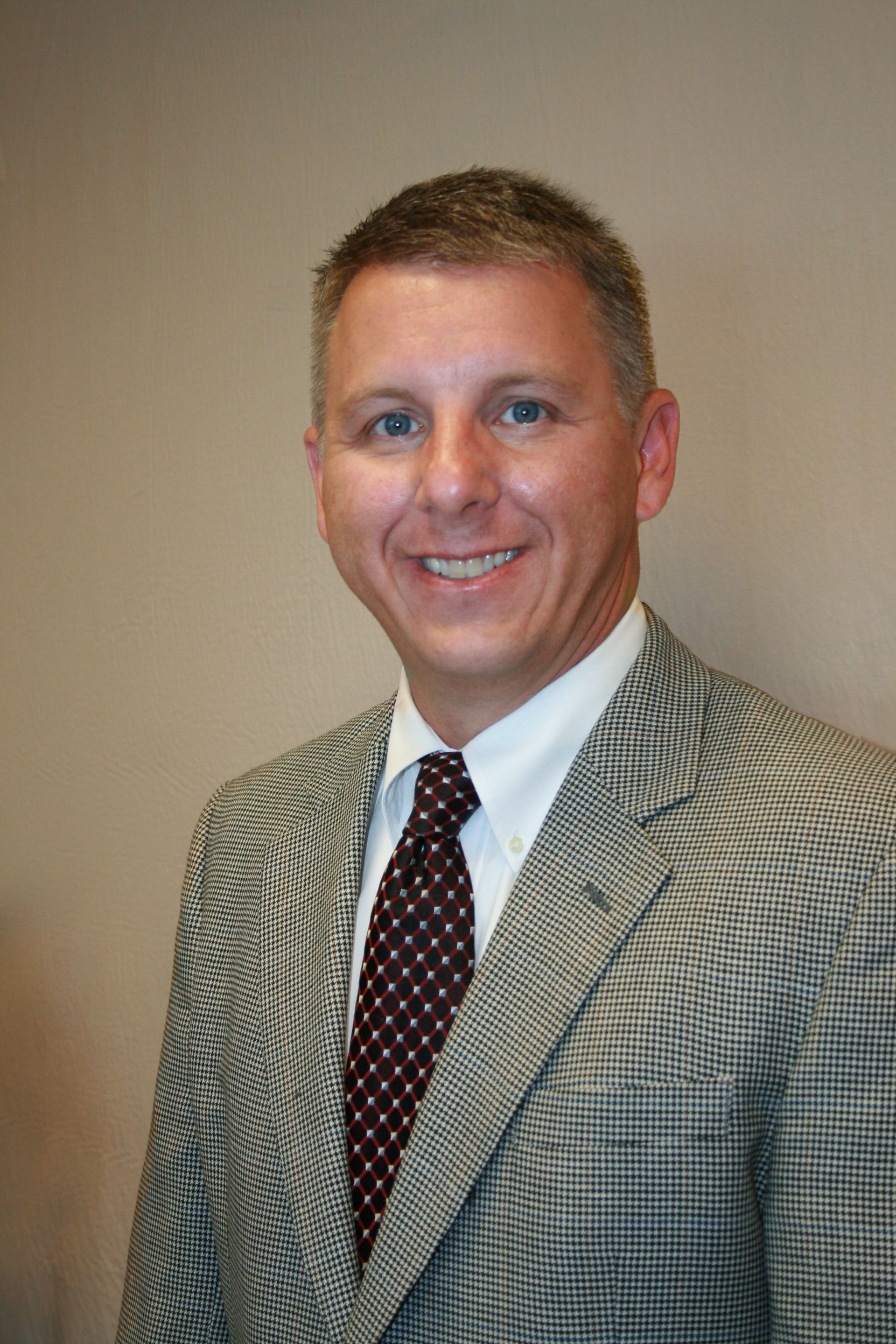 A person in a patterned blazer, white dress shirt, and a dark patterned tie, smiling against a plain light-colored wall.