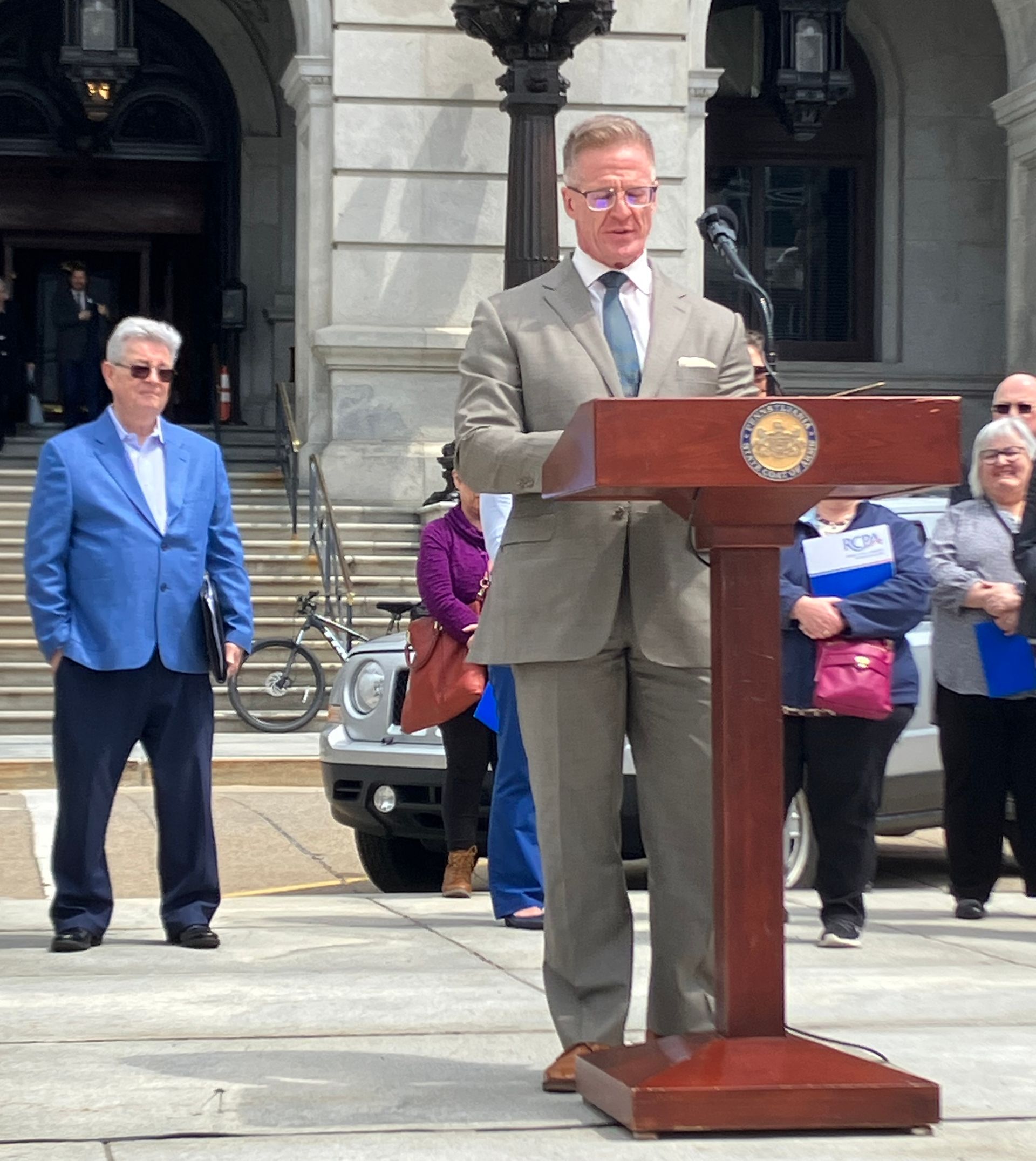 A person in a tan suit stands at a wooden podium outdoors, speaking to a group of people near a stone building.