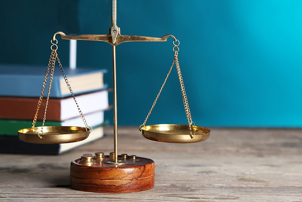 A brass scale of justice sits on a wooden surface with a stack of colorful books in the background.