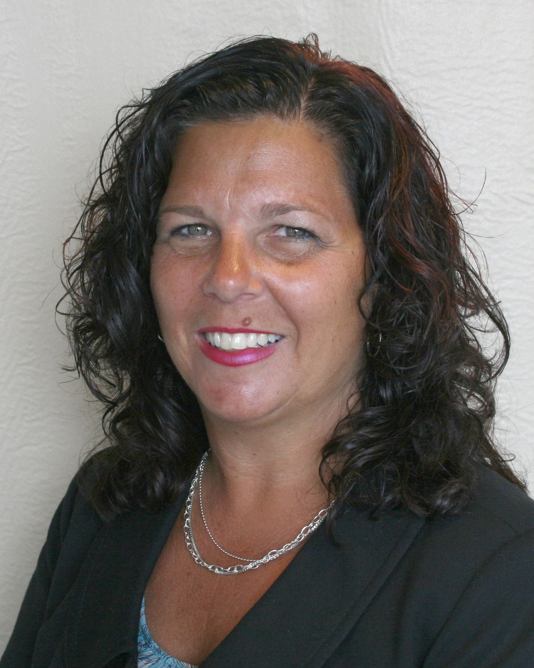 A professional headshot of a smiling person with dark, wavy hair, wearing a black blazer and a silver chain necklace.