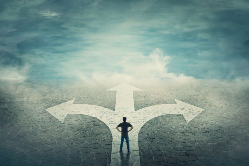 A person stands before a fork in the road with three diverging arrows painted on a textured ground under a cloudy sky.