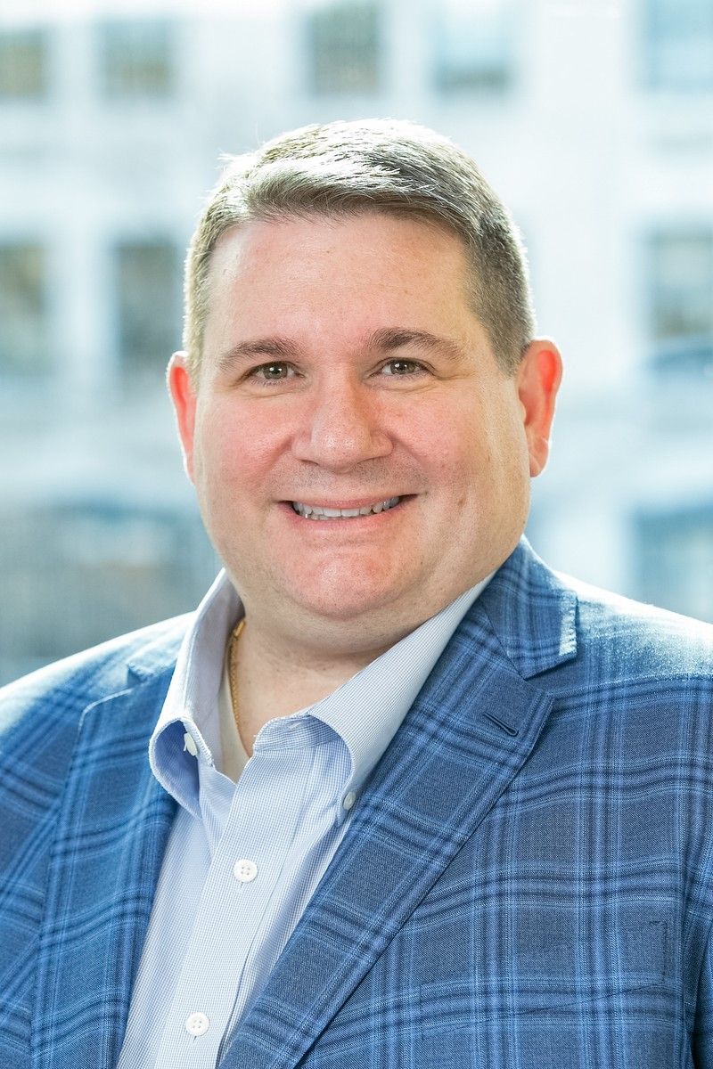 Smiling man in a blue plaid blazer and light shirt against a blurred office background