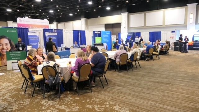 People seated at round tables in a conference hall with blue exhibit booths and a large screen.