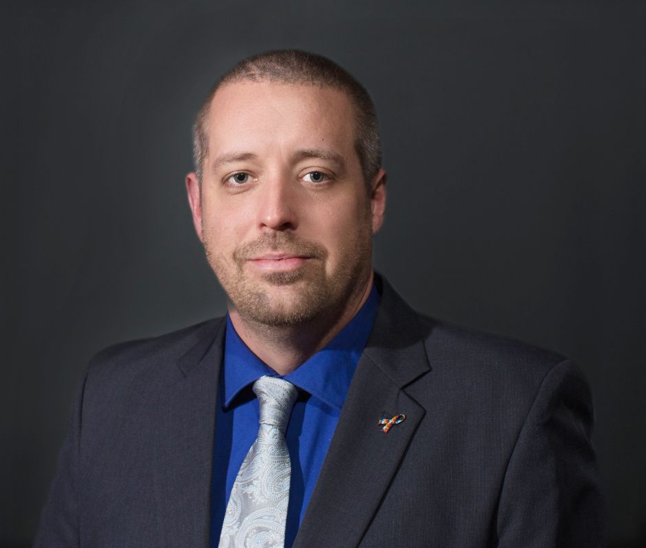 A person in a charcoal suit, blue shirt, and light-patterned tie poses against a dark gray background.