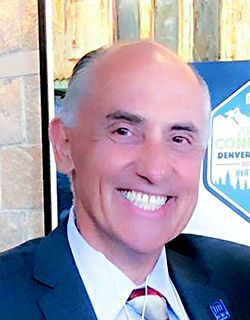 Keynote Presenter Name Smiling man in a suit and tie standing indoors at an event backdrop