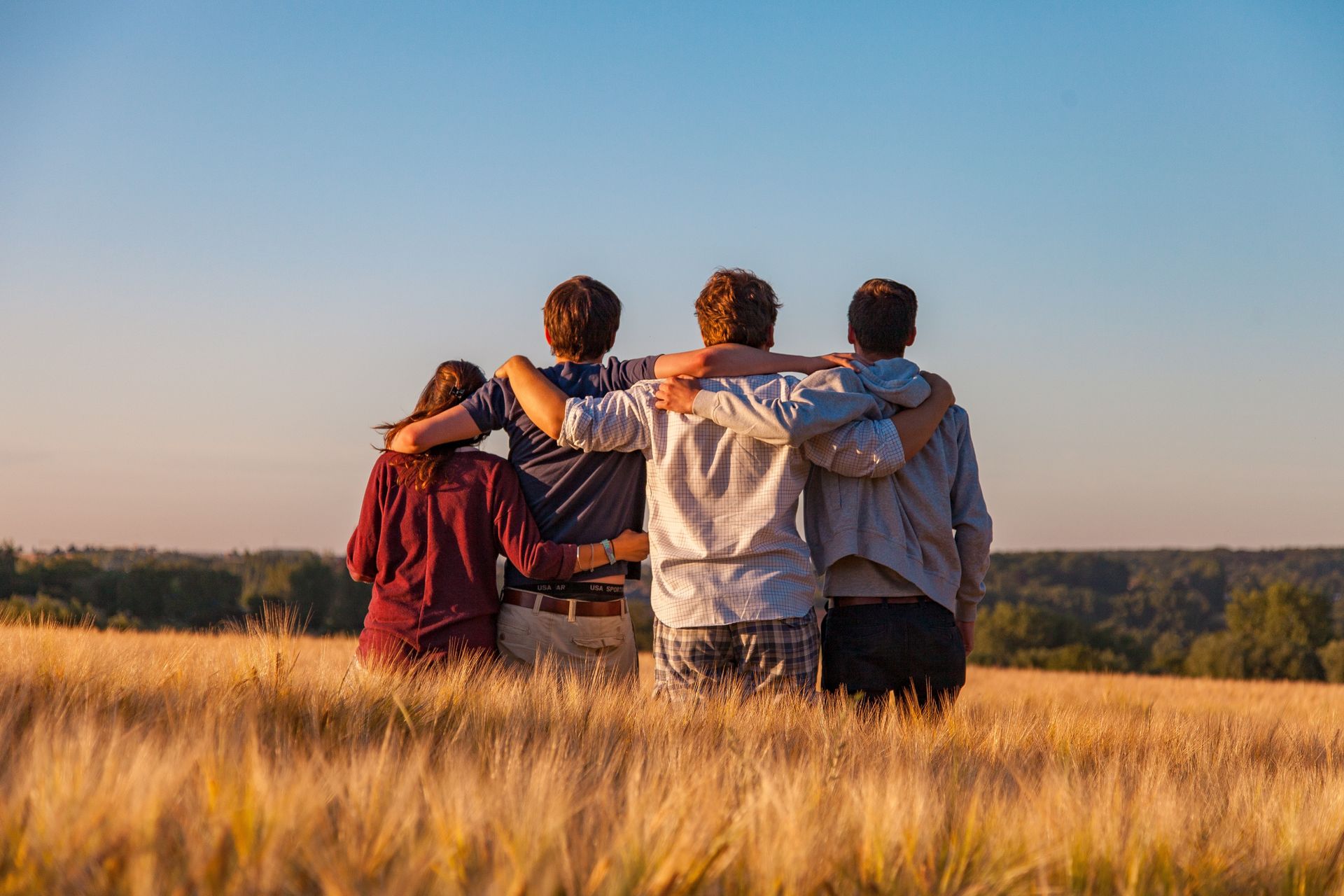 Four individuals in a field, with arms over each other's shoulders in support, looking away from the