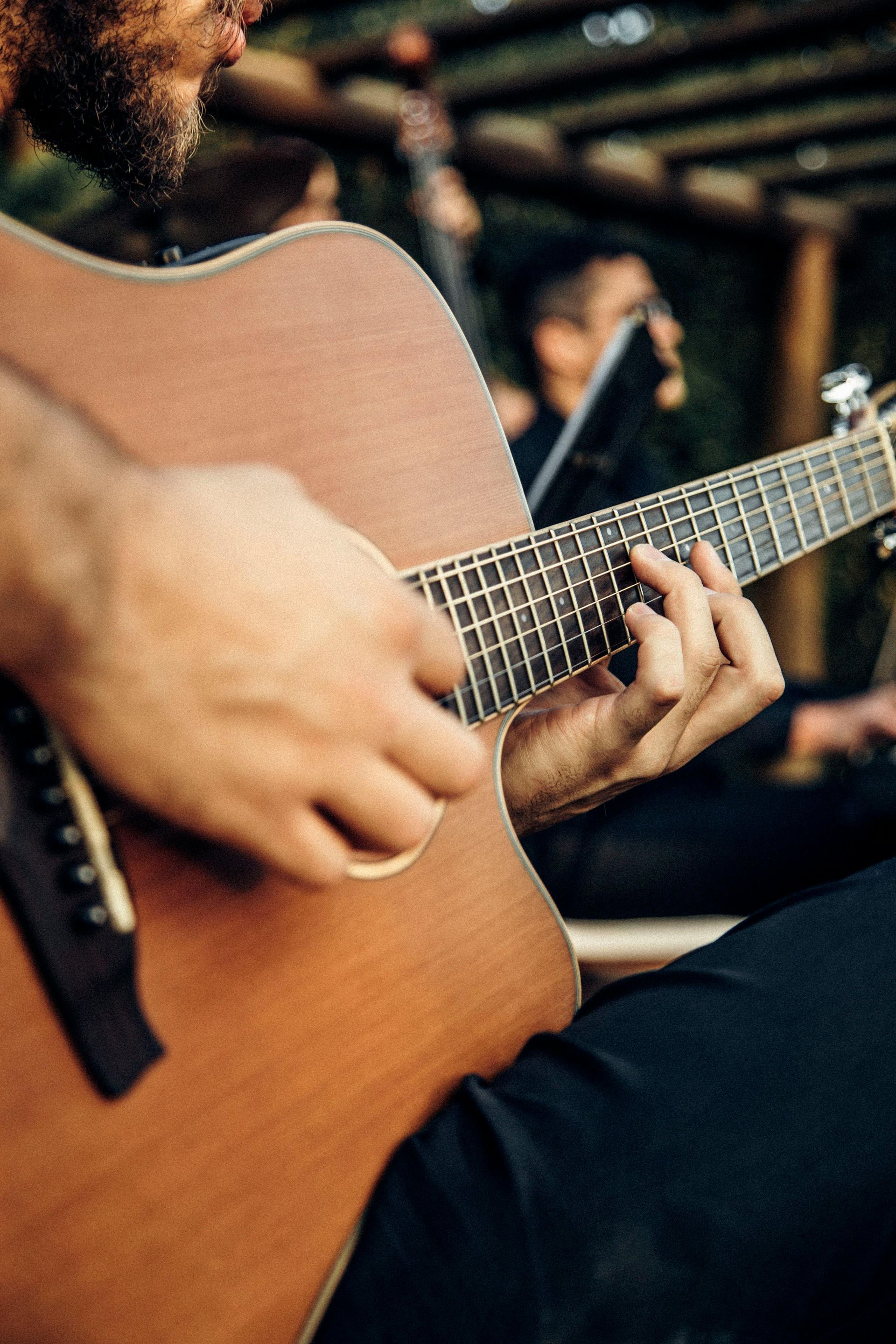 Close-up of hands playing an acoustic guitar, with a blurred band member in the background