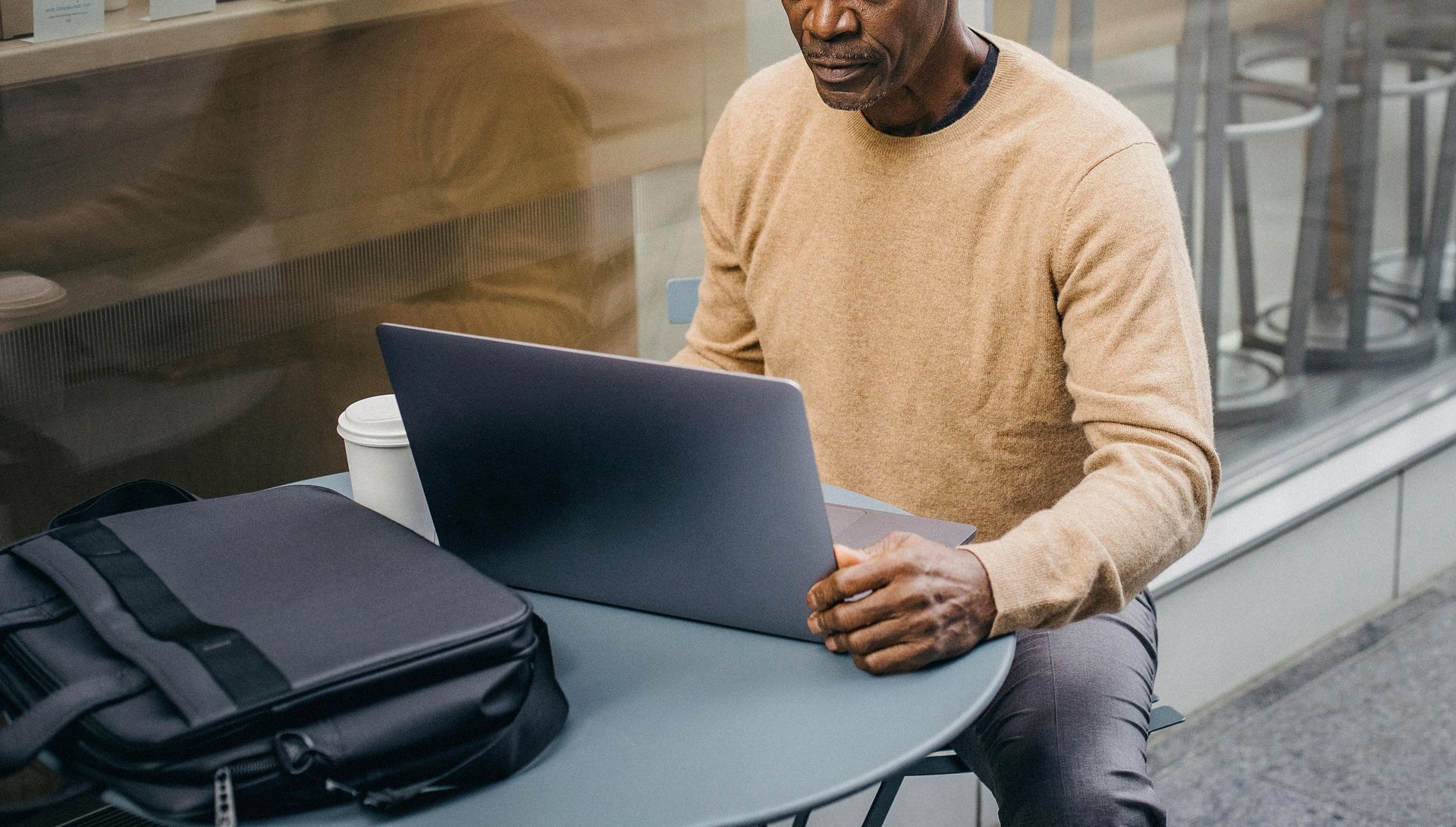 A person in a beige sweater sits at a round outdoor table using a laptop next to a black messenger bag.