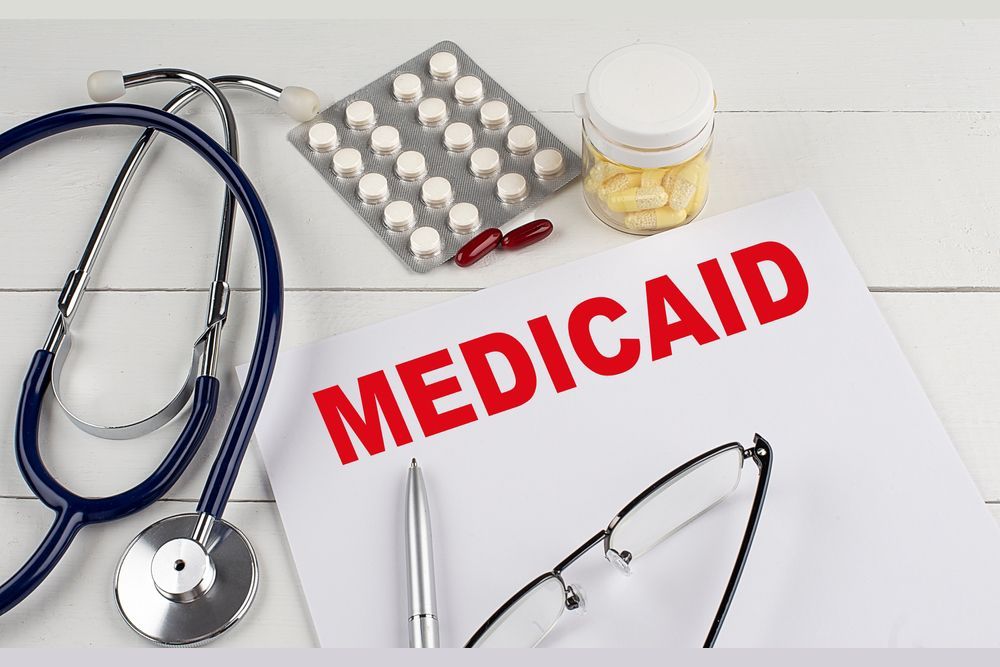 MEDICAID written in thick, red font on paper, surrounded by medicine tablets and a stethoscope