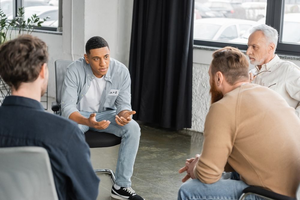 A group of people sit in a circle in a bright office, listening to a person gesturing while speaking during a meeting.