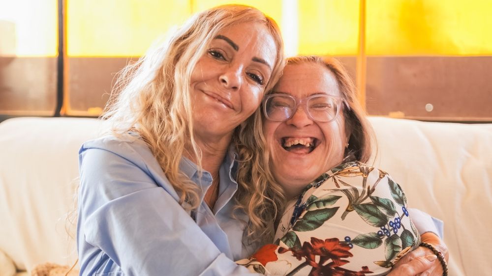 Two smiling women hugging on a couch in warm sunlight