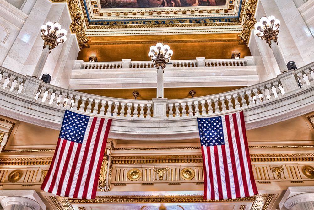 Low-angle view of two American flags hanging in a grand, ornate rotunda with classic columns and gold architectural trim.