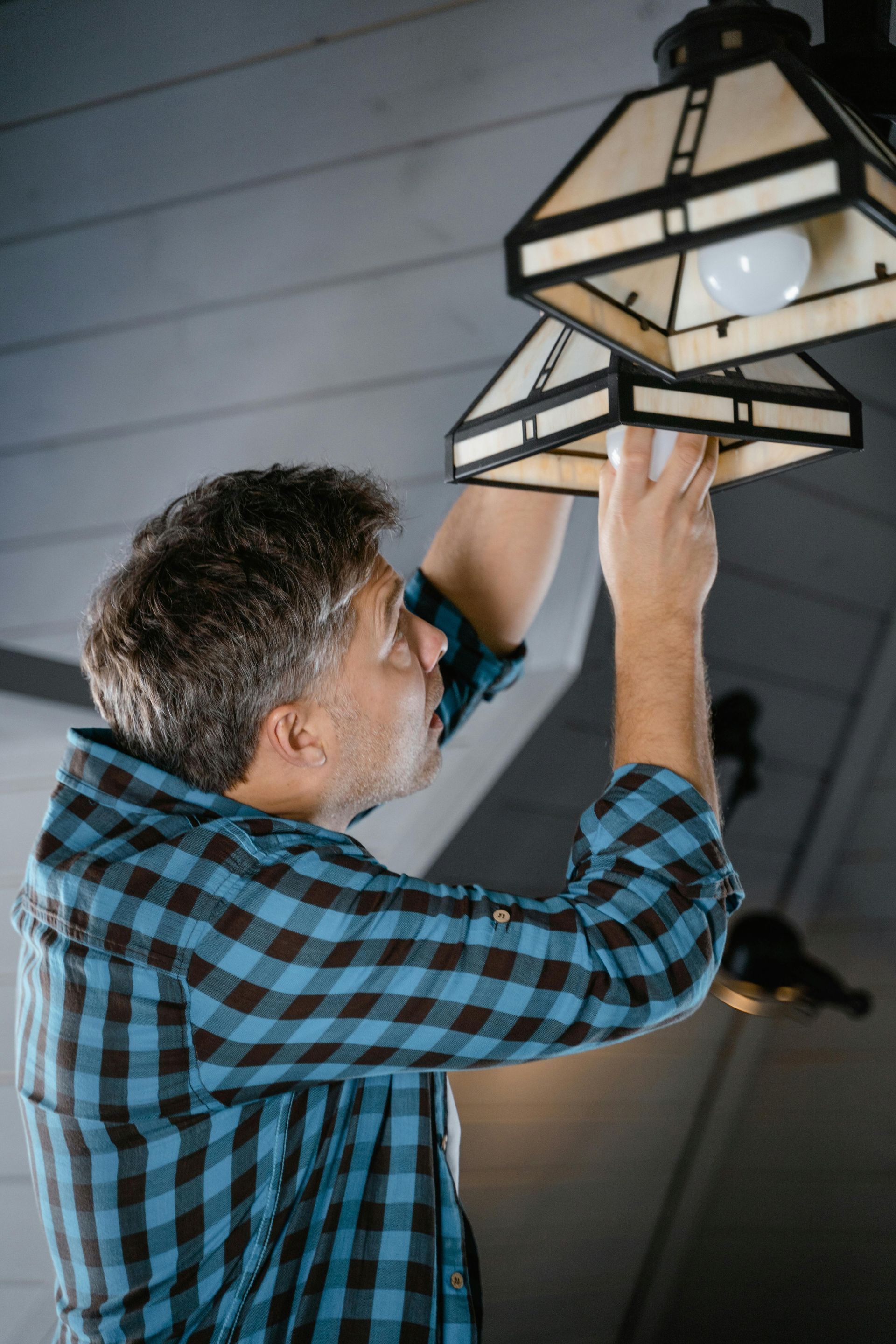 Man in plaid shirt replacing a lightbulb in a hanging light fixture. Indoors, gray ceiling.