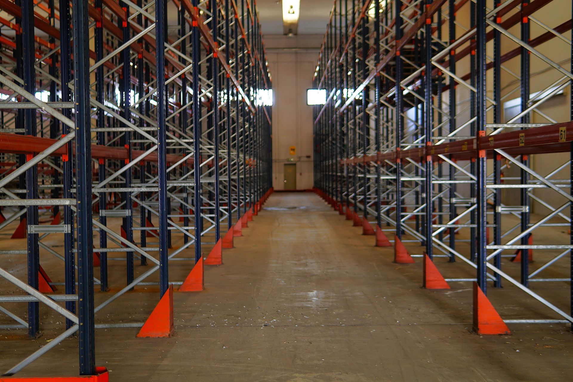 Allée d'entrepôt vide bordée de part et d'autre de hautes étagères industrielles métalliques bleues et oranges.