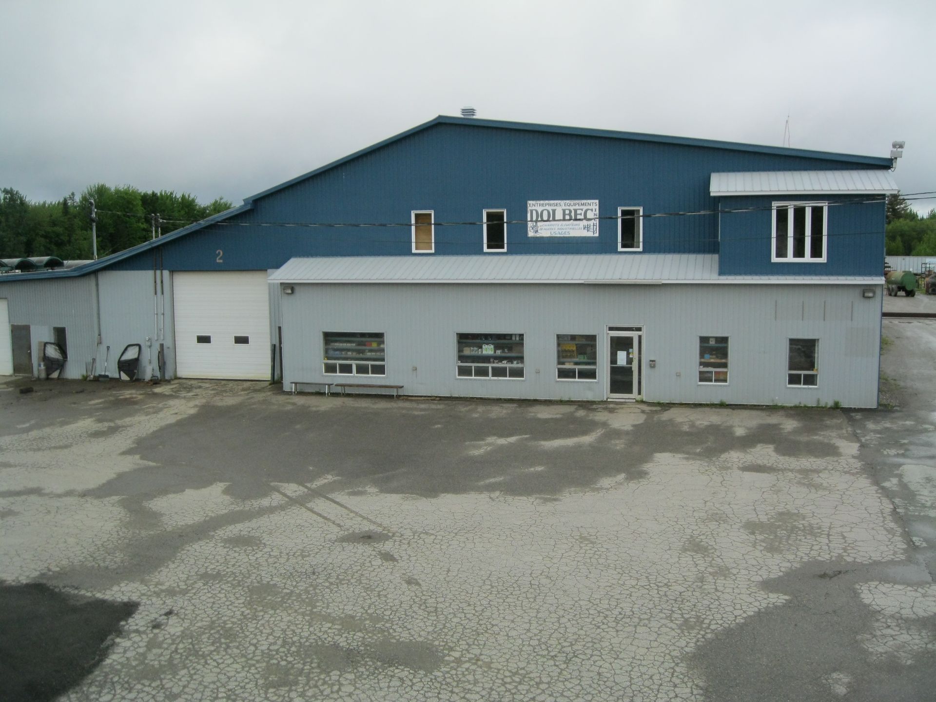 Un bâtiment commercial bleu et gris avec une porte de garage blanche, de petites fenêtres et un parking pavé à l'avant.